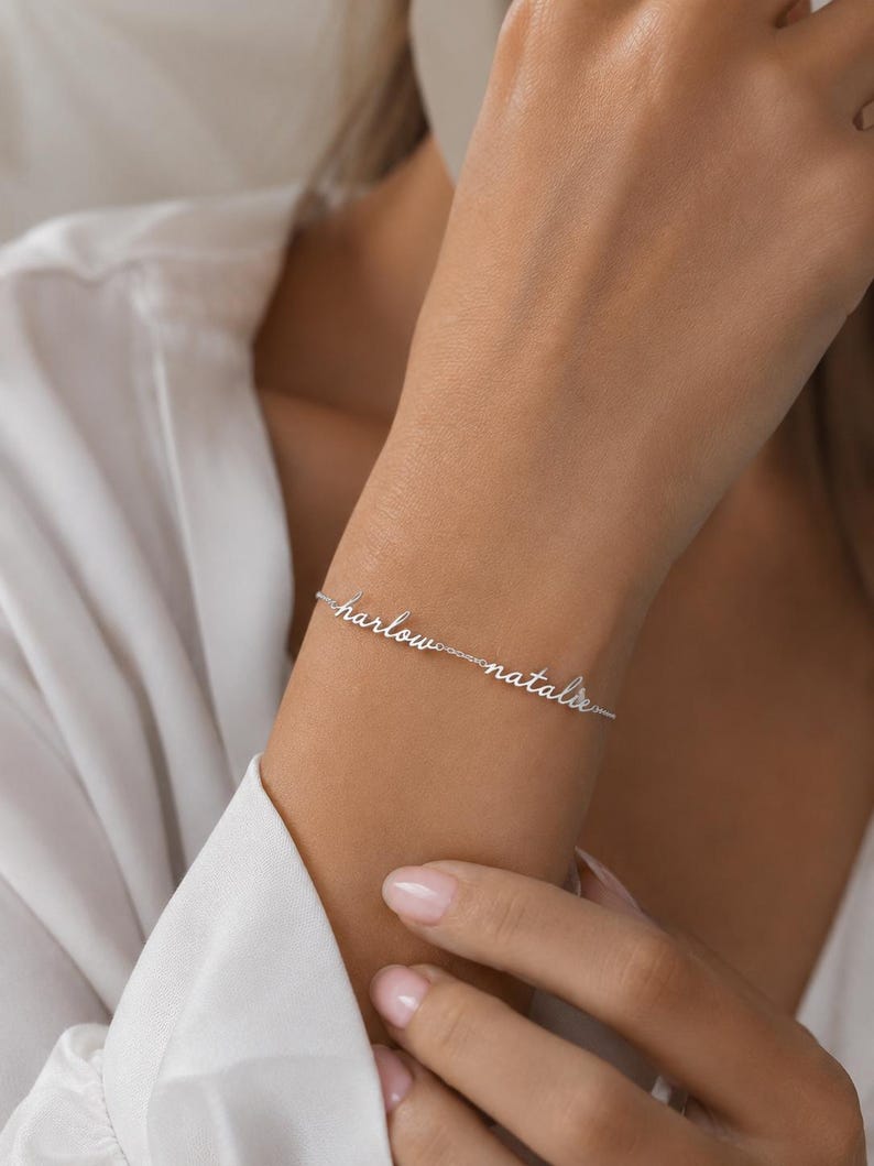 K&ouml;nnte beinhalten: Ein silberfarbenes Kettenarmband mit den zwei Namen Harlow und Natalie in kursiver Schreibschrift. Die Namen sind durch eine feine Metallkette miteinander verbunden und bilden ein durchgehendes Schmuckst&uuml;ck auf der Haut.