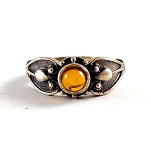 May include: A silver ring with an amber-colored stone at the center. The ring has an ornate design with leaf-like shapes and small, round accents on either side of the stone. The ring is set against a white background.