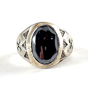 May include: A silver-toned ring featuring a large, oval-shaped, faceted black gemstone set in the center. The metal band displays a textured, engraved geometric pattern along the sides, providing a detailed contrast to the smooth, dark stone.