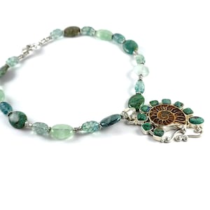 May include: A green and blue gemstone necklace with a silver pendant featuring a fossilized ammonite surrounded by turquoise stones.