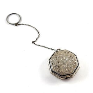 May include: A silver-colored, octagonal tea caddy with intricate floral engravings. It features a small ring and a chain attached to the caddy. The caddy is likely made of metal and is displayed against a white background.