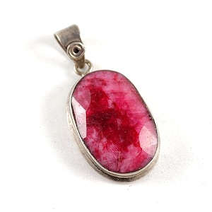 May include: A silver pendant with a large, oval-shaped ruby gemstone. The ruby is a deep red color with visible inclusions and facets. The pendant has a simple, elegant design, suitable for everyday wear or special occasions.