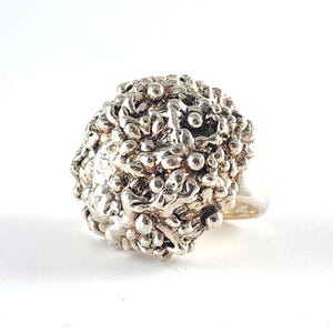 May include: A silver ring featuring a large, rounded top section with a highly textured, organic surface composed of numerous small, raised metallic spheres and irregular molten-like shapes that create a dense, clustered, and reflective appearance.