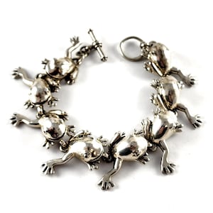May include: A silver-toned bracelet featuring a series of frog figurines. Each frog is detailed with outstretched limbs and a textured surface. The bracelet has a toggle clasp closure. The frogs are arranged in a circular pattern.