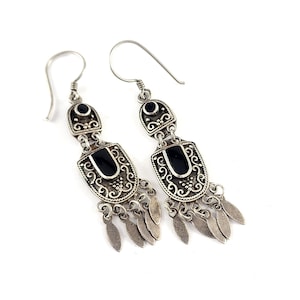 May include: A pair of silver-toned dangle earrings. Each earring features a rectangular black stone set in an ornate, filigree design. Small, leaf-shaped charms hang from the bottom. The earrings have a hook closure.