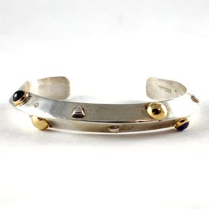 May include: Silver cuff bracelet with gold and black accents. The bracelet features a curved design with gold studs and black gemstones. The metal is polished and the bracelet is open at the ends.