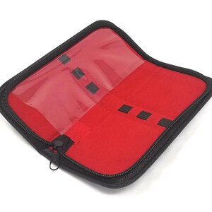 Pode incluir: Uma caixa preta com zíper, com um interior vermelho e múltiplos slots para guardar pequenos objetos. A caixa tem uma janela de plástico transparente na frente.