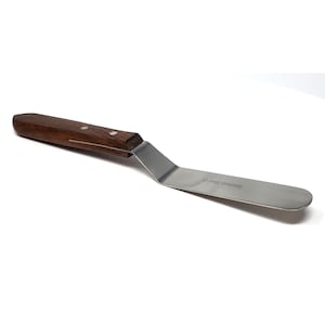 May include: An offset spatula with a dark brown wooden handle secured by two metal rivets and a flat, flexible stainless steel blade. The metal blade features an angled neck and is clearly stamped with the text Stainless Steel CE.