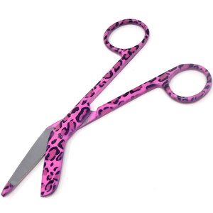 May include: Pink bandage scissors with a leopard print design. The scissors have a curved blade and a sharp point.