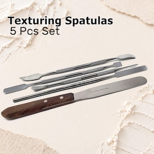 May include: A 5-piece set of texturing spatulas featuring four double-ended stainless steel tools with textured grips and one larger spatula with a wooden handle. The text "Texturing Spatulas 5 Pcs Set" is printed in black above the metal tools.
