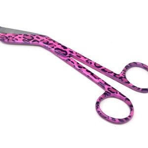 May include: Pink bandage scissors with a leopard print design. The scissors have a curved blade and a ring handle.