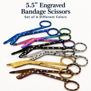 Op de afbeelding: Een set van zes 14 cm gegraveerde verbandscharen in diverse kleuren, waaronder blauw, regenboog, zwart, zilver, goud en bruin. Elke schaar is voorzien van een herhalend patroon van pootafdrukken gegraveerd op de bladen en de handvatten.