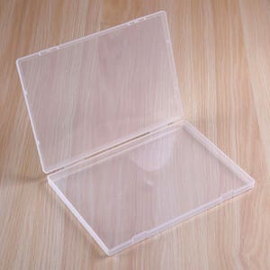 May include: A rectangular, clear plastic storage case with a hinged lid. The case features a smooth, semi-transparent finish and a simple latch mechanism along the front edge, designed for organizing small items or documents in a flat, open position.