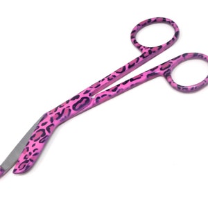 May include: Pink bandage scissors with a leopard print design. The scissors have a curved blade and are made of stainless steel.
