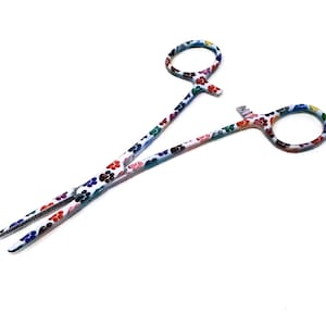 Puede incluir: Un par de pinzas hemostáticas blancas con un diseño de huella de pata de colores. Las pinzas hemostáticas están cerradas y tienen una punta dentada.