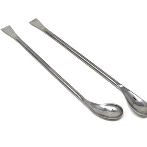 May include: Two stainless steel laboratory spatulas with rounded ends. The spatulas are approximately 25 cm long.