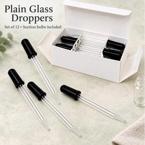 May include: A set of 12 clear glass droppers with black rubber suction bulbs, presented in a white cardboard box. The text Plain Glass Droppers Set of 12 Suction bulbs included is visible. Four individual droppers are arranged in front.