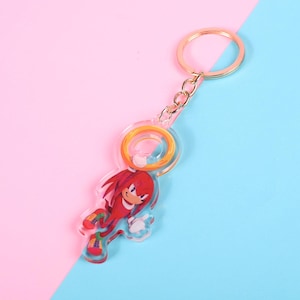 May include: A clear acrylic keychain featuring a red cartoon character with white gloves and green and red shoes. The keychain has a gold-colored ring and chain. The background is split between pink and blue.