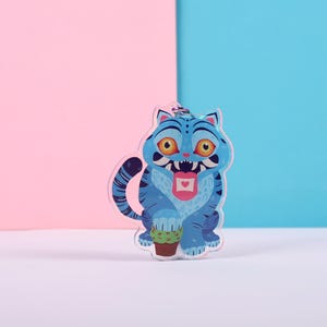 Puede incluir: Un llavero azul con forma de gato con un diseño de dibujos animados. El gato tiene la boca muy abierta con una lengua rosa y un pequeño corazón. Sostiene una pequeña planta en maceta. El fondo es rosa y azul.