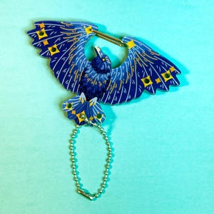 May include: A blue bird-shaped keyring with yellow and green details. The bird has outstretched wings and a silver chain attached. The keyring is set against a light blue backdrop.