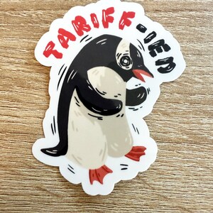 May include: A white sticker with a cartoon penguin wearing a red beanie and holding a red cup. The text "TARIFF-FIED" is written in red around the penguin.