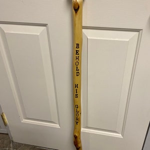 Op de afbeelding: Een houten wandelstok met een bruine, bolvormige bovenkant en een grijze rubberen punt. De woorden "BEHOLD HIS GLORY" staan verticaal in zwart op het lichtgekleurde hout. De stok heeft een natuurlijk, rustiek uiterlijk.
