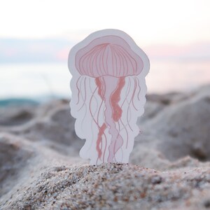 Puede incluir: Un recorte de papel rosa y blanco de una medusa en una playa de arena.