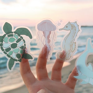 Puede incluir: Cuatro pegatinas transparentes con dise&ntilde;os de vida marina. Una tortuga marina verde, una medusa rosa, un caballito de mar azul claro y un delf&iacute;n azul claro.