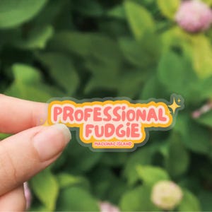 Könnte beinhalten: Ein transparenter Aufkleber mit dem Text "PROFESSIONAL FUDGIE" in Rosa mit gelbem Rand. Darunter steht "MACKINAC ISLAND". Auf der rechten Seite des Aufklebers befindet sich ein kleiner gelber Stern.