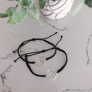 May include: Two black string bracelets with silver wave charms. The bracelets are tied together.
