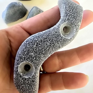 May include: Gray climbing holds with a textured surface and pre-drilled holes. The holds are in various shapes, including a curved, wave-like design. Several other gray climbing holds are visible in the background. These holds are designed for rock climbing.