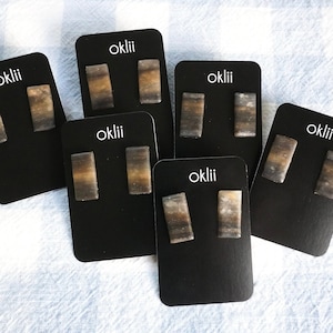May include: Six pairs of earrings on black cards with the word "oklii" printed on each card. The earrings are rectangular and have a brown, white, and gray marbled pattern.
