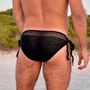 Puede incluir: Braga de baño negra de ganchillo con un diseño similar a una red. La braga tiene cierres de lazo a cada lado. La imagen está tomada en una playa con un fondo borroso de follaje verde y cielo azul claro.