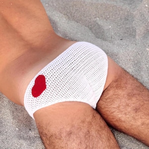 Puede incluir: Ropa de baño blanca de ganchillo con un motivo de corazón rojo. El material de punto abierto crea una apariencia texturizada. El corazón es de un rojo intenso. Esta ropa de baño es adecuada para la playa o la piscina.