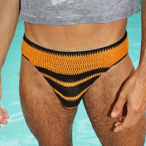 Puede incluir: Braguita de traje de baño de ganchillo hecha a mano con un patrón de rayas negras y amarillo dorado. El traje de baño está diseñado con una cintura baja y un corte de calzón. La imagen está tomada al aire libre con un fondo de agua azul.