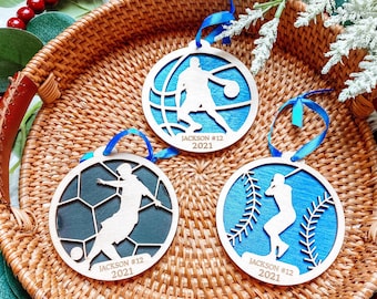 May include: Three wooden ornaments with blue, white, and black paint. Each ornament features a silhouette of a person playing a different sport: basketball, soccer, and baseball. The ornaments are personalized with the name "Jackson #12 2021".