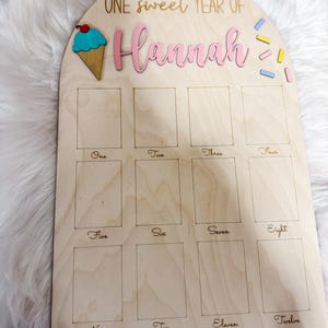 May include: Wooden milestone board with the text "ONE sweet YEAR OF Hannah". The board features an ice cream cone and colorful sprinkles. It has twelve rectangular spaces labeled with the numbers one through twelve.