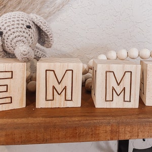 Puede incluir: Bloques de madera que deletrean el nombre "EMMA" en may&uacute;sculas. Los bloques son de color marr&oacute;n claro y las letras est&aacute;n talladas en la madera. Un peque&ntilde;o juguete de elefante de crochet est&aacute; sentado en la parte superior del primer bloque.