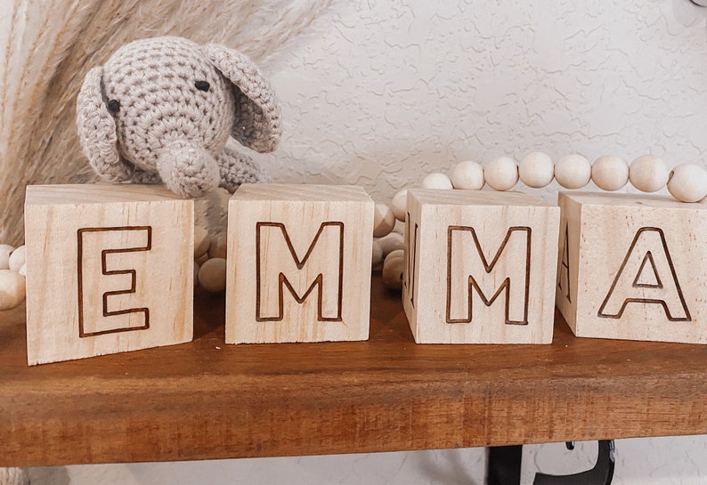 Puede incluir: Bloques de madera que deletrean el nombre "EMMA" en may&uacute;sculas. Los bloques son de color marr&oacute;n claro y las letras est&aacute;n talladas en la madera. Un peque&ntilde;o juguete de elefante de crochet est&aacute; sentado en la parte superior del primer bloque.