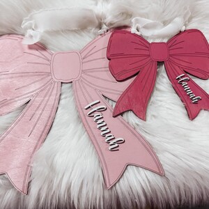May include: Two pink and red wooden bow ornaments with the name "Hannah" written on each. The bows are on a white fluffy surface.