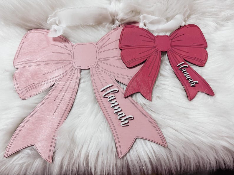 May include: Two pink and red wooden bow ornaments with the name "Hannah" written on each. The bows are on a white fluffy surface.