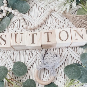 Puede incluir: Bloques de madera que deletrean el nombre "Sutton" sobre un fondo blanco de crochet con hojas verdes y un mordedor de madera.