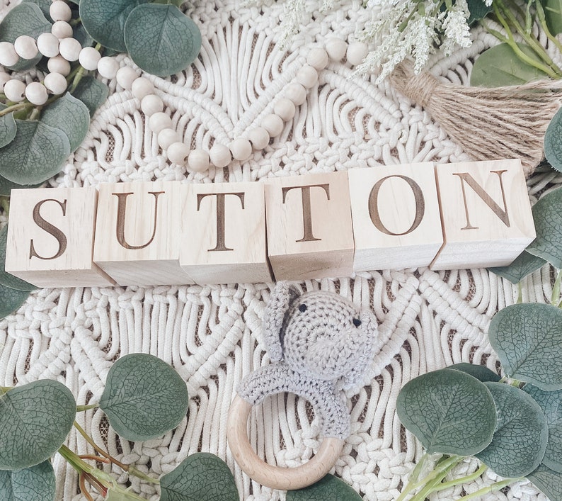 Puede incluir: Bloques de madera que deletrean el nombre "Sutton" sobre un fondo blanco de crochet con hojas verdes y un mordedor de madera.
