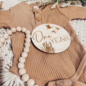 May include: A wooden round sign with the text "One Year" and a design of flying birds and reeds. The sign is on top of a brown ribbed onesie with a wooden bead necklace.