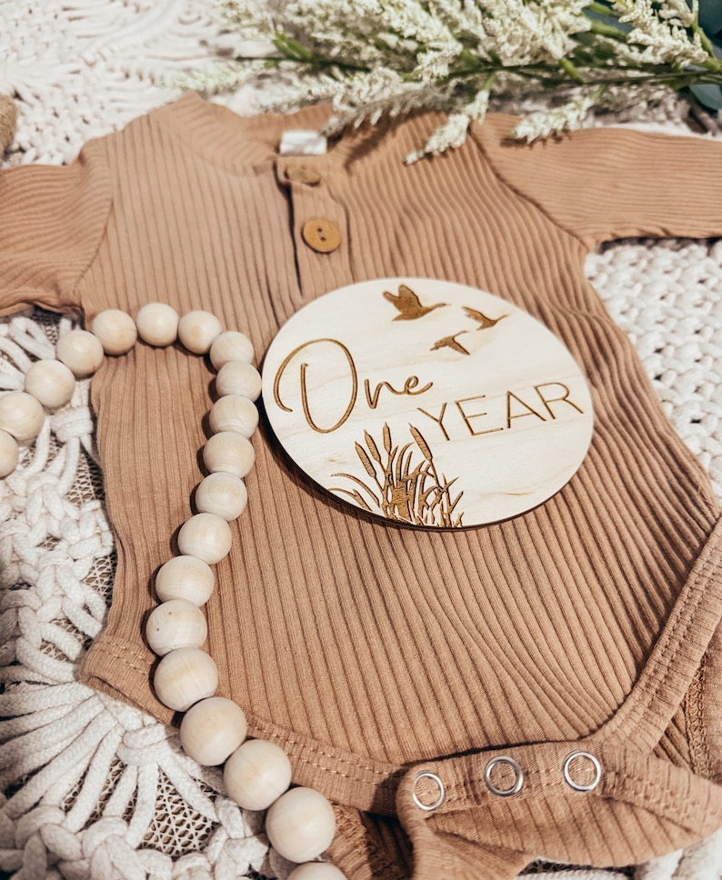 May include: A wooden round sign with the text "One Year" and a design of flying birds and reeds. The sign is on top of a brown ribbed onesie with a wooden bead necklace.