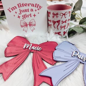 May include: Two wooden bow ornaments with the names "Macie" and "Paisley" written on them. The bows are pink and purple and are on a white fluffy surface.