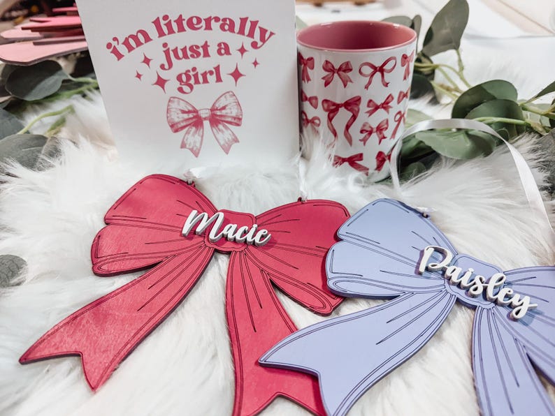 May include: Two wooden bow ornaments with the names "Macie" and "Paisley" written on them. The bows are pink and purple and are on a white fluffy surface.