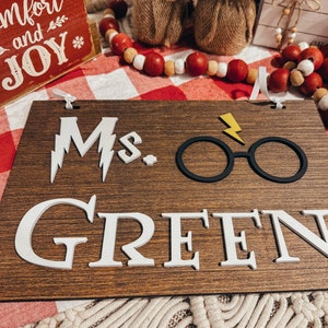 May include: A wooden sign with the text "Ms. Green" in white letters. The sign also features a black lightning bolt and a pair of black glasses.