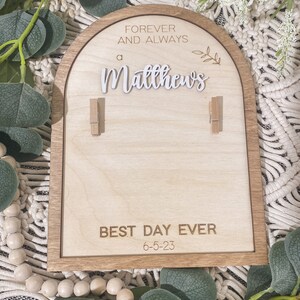 Może przedstawiać: Drewniany znak z zaokrąglonym wierzchołkiem i brązową ramą. Znak głosi "Forever and Always a Matthews" z małym wzorem gałęzi. M&oacute;wi r&oacute;wnież "Best Day Ever 6-5-23" i ma dwa drewniane spinacze.