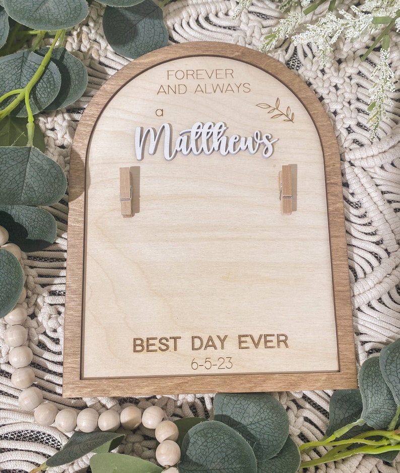 Może przedstawiać: Drewniany znak z zaokrąglonym wierzchołkiem i brązową ramą. Znak głosi "Forever and Always a Matthews" z małym wzorem gałęzi. M&oacute;wi r&oacute;wnież "Best Day Ever 6-5-23" i ma dwa drewniane spinacze.